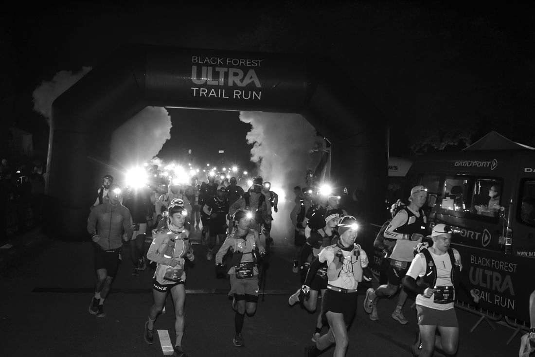 Nachtstart 2026: Läufer*innen mit Stirnlampen laufen durch Nebel und Licht aus dem Startbogen